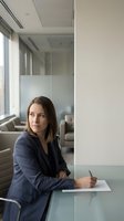 Professional businesswoman in navy suit writing at glass conference table in modern corporate office with natural lighting.