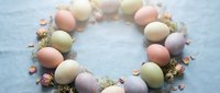 Pastel Easter eggs arranged in a wreath circle with dried flowers on soft blue linen background, spring holiday flat lay composition.