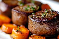 Grilled beef tenderloin medallions with roasted carrots, garnished with fresh herbs and seasonings, served on white plate. Close up view shows juicy meat texture.