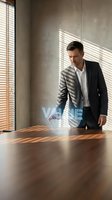 Professional businessman in dark suit standing at wooden conference table in modern office with natural sunlight streaming through venetian blinds.