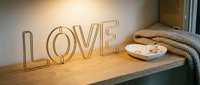 Gold wire LOVE sign glowing warmly on a wooden shelf beside a cozy knit blanket and small decorative dish in a modern interior.