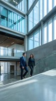 Two business professionals in suits walking down stairs in a modern glass corporate building, discussing strategy.