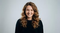 Young woman with curly brown hair smiling warmly in black sweater against neutral background for professional headshot and portrait photography.