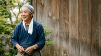 Senior Asian man with gray hair wearing navy blue cardigan and white undershirt, smiling outdoors against weathered wooden wall with green foliage, portrait for lifestyle and wellness content.