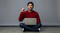 Young South Asian man in red sweater sitting cross-legged with laptop pointing finger up having eureka moment against gray background for business concepts.