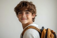Young Caucasian teen boy with curly brown hair wearing white sweater and yellow backpack smiling at camera against light background. Natural daylight portrait.