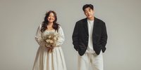 Young Asian bride in elegant white wedding dress holding bouquet stands beside groom in black suit jacket and white pants against neutral studio background.