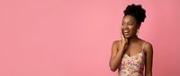 Young African American woman with natural curly hair laughing joyfully in colorful floral dress against pink studio background for lifestyle content.