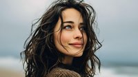 Young woman with windswept dark hair smiling at seaside, wearing brown sweater against blurred ocean background. Natural beauty portrait for lifestyle content.