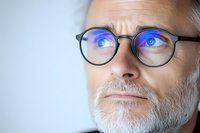 Middle-aged Caucasian man with gray beard and round glasses reflecting blue light, looking thoughtfully aside against light background. Digital eye strain concept.