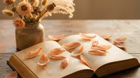 Autumn reading scene with open book, fallen orange leaves and dried flowers in rustic vase on wooden table creating cozy seasonal atmosphere for relaxation.
