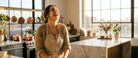 Happy young woman laughing in modern kitchen with natural sunlight streaming through large windows creating warm atmosphere for cooking and lifestyle content.