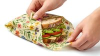 Hands wrapping healthy sandwich with vegetables in eco-friendly reusable beeswax food wrap with floral pattern for sustainable lunch storage.