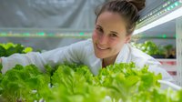 Young Caucasian female farmer tending to fresh lettuce in modern hydroponic greenhouse system, wearing white coat and smiling while checking leafy greens growth.