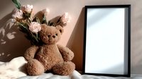 Cute brown teddy bear sitting next to pink roses and blank picture frame on wooden surface with soft natural lighting for nursery decor and gift concepts.
