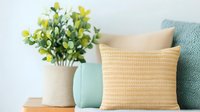 Cozy home decor arrangement with soft textured pillows in mint green and beige tones, complemented by artificial yellow flowers in ceramic vase against white wall.
