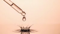 Cosmetic serum dropper releasing golden oil drop creating elegant water splash crown effect on soft pink background for skincare beauty products.