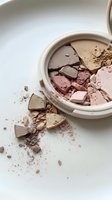 Broken makeup powder compact with scattered cosmetic debris on white surface for beauty and fashion concepts.