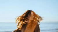 Young woman with flowing wavy hair moving in wind against ocean backdrop during golden hour sunset creating dreamy atmospheric portrait for lifestyle content.