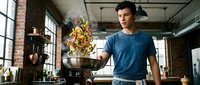 Young Caucasian male chef tossing colorful vegetables in wok pan at modern industrial kitchen. Professional cooking technique demonstration in loft-style restaurant.