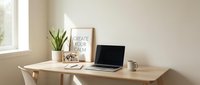 Minimalist home office workspace with laptop, motivational poster, green plant, and mug on a wooden desk in a bright, neutral room.