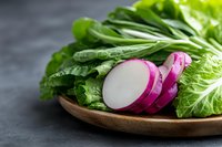 Fresh sliced red radish and green lettuce leaves arranged on wooden plate against dark background, healthy organic vegetables for salad preparation.