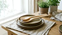 Wooden salad servers resting on stacked ceramic plates with linen napkin on oak table, natural kitchen decor with green herbs in background.