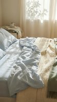 Unmade bed with white linens, cream blanket, and beige throw in bright morning sunlight from window with sheer curtains, cozy bedroom interior design.