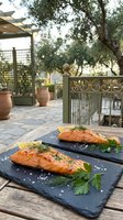 Grilled salmon fillets with herbs on slate plates served on outdoor terrace table for Mediterranean dining experience.