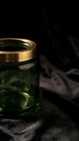 Glass jar with gold lid containing green liquid on dark fabric background, suitable for cosmetics, skincare, or luxury product packaging design.