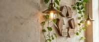 Rustic wooden recycling symbol on a textured stone wall with vintage pendant lights and trailing green ivy vines in warm light.