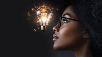 Young African American woman in glasses gazing at glowing light bulb with magical sparkles and energy, symbolizing innovation, creativity and bright ideas.