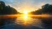 Tranquil river landscape at sunrise with golden sunlight breaking through morning mist over calm water surface, trees silhouetted against dramatic dawn sky.