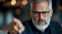 Confident mature businessman with gray hair and beard wearing glasses points forward against blurred bokeh background, showing leadership and authority.