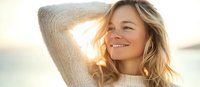 Young Caucasian woman with blonde wavy hair smiling at sunset, wearing cream sweater. Natural backlit portrait with warm golden light and peaceful expression.