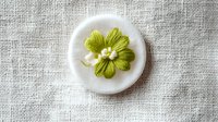 Delicate lime green embroidered flower on white ceramic button against natural linen fabric background, showcasing intricate needlework detail and craftsmanship.