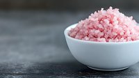 Pink Himalayan salt crystals in white ceramic bowl on dark stone background, macro view of gourmet mineral seasoning for cooking and spa treatments.