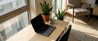 Modern home office workspace with laptop computer on wooden desk near window with natural sunlight and green plants creating productive work environment.