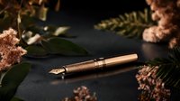 Elegant copper-colored fountain pen on dark background with soft pink dried flowers and green leaves creating luxurious composition for stationery and writing themes.