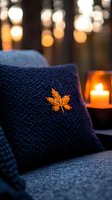 Cozy navy knitted pillow with golden maple leaf embroidery against warm candlelight bokeh background creates peaceful autumn evening atmosphere.