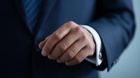 Close-up of businessman's hand in formal navy suit with white shirt cuff visible against blue background, conveying professional confidence in corporate setting.
