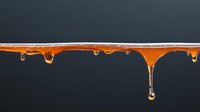 Amber honey drips and forms translucent drops against dark background, creating natural abstract pattern with golden liquid strands and falling droplets.