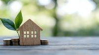 Wooden house model with green plant and coins on table representing eco-friendly real estate investment and sustainable home financing growth concept.