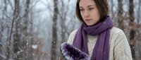 Young Caucasian woman in purple scarf and cream sweater holding snow in winter forest with falling snowflakes creating peaceful seasonal moment.