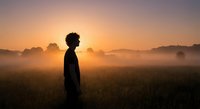 Young African American man silhouette standing in misty field during golden sunrise with warm atmospheric lighting and peaceful countryside landscape.