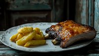 Grilled pork ribs with golden roasted potatoes served on vintage plate in rustic kitchen setting for culinary and food photography projects.