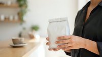 Woman in black blazer holding white kraft paper bag with packaging mockup on wooden table in modern office setting.