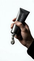 Male hand holding black grooming product tube with gray paste dispensed, isolated on white background for men's skincare and cosmetics advertising.