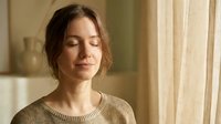 Young woman with closed eyes enjoying peaceful moment in warm sunlight by window at home for wellness and mindfulness meditation practices.