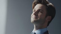 Young Caucasian businessman with confident smile looking up dreamily near window, wearing blue suit. Natural light creates atmospheric mood for aspirational corporate portrait.